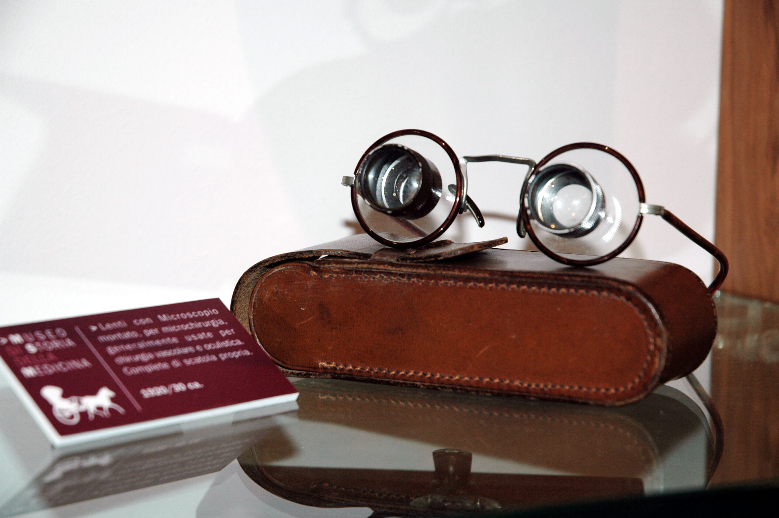Eine runde Vintage-Brille mit dicken Gläsern liegt auf einem braunen Lederetui, das auf einer Glasfläche mit einem unscharfen Informationsschild in der Nähe ausgestellt ist.