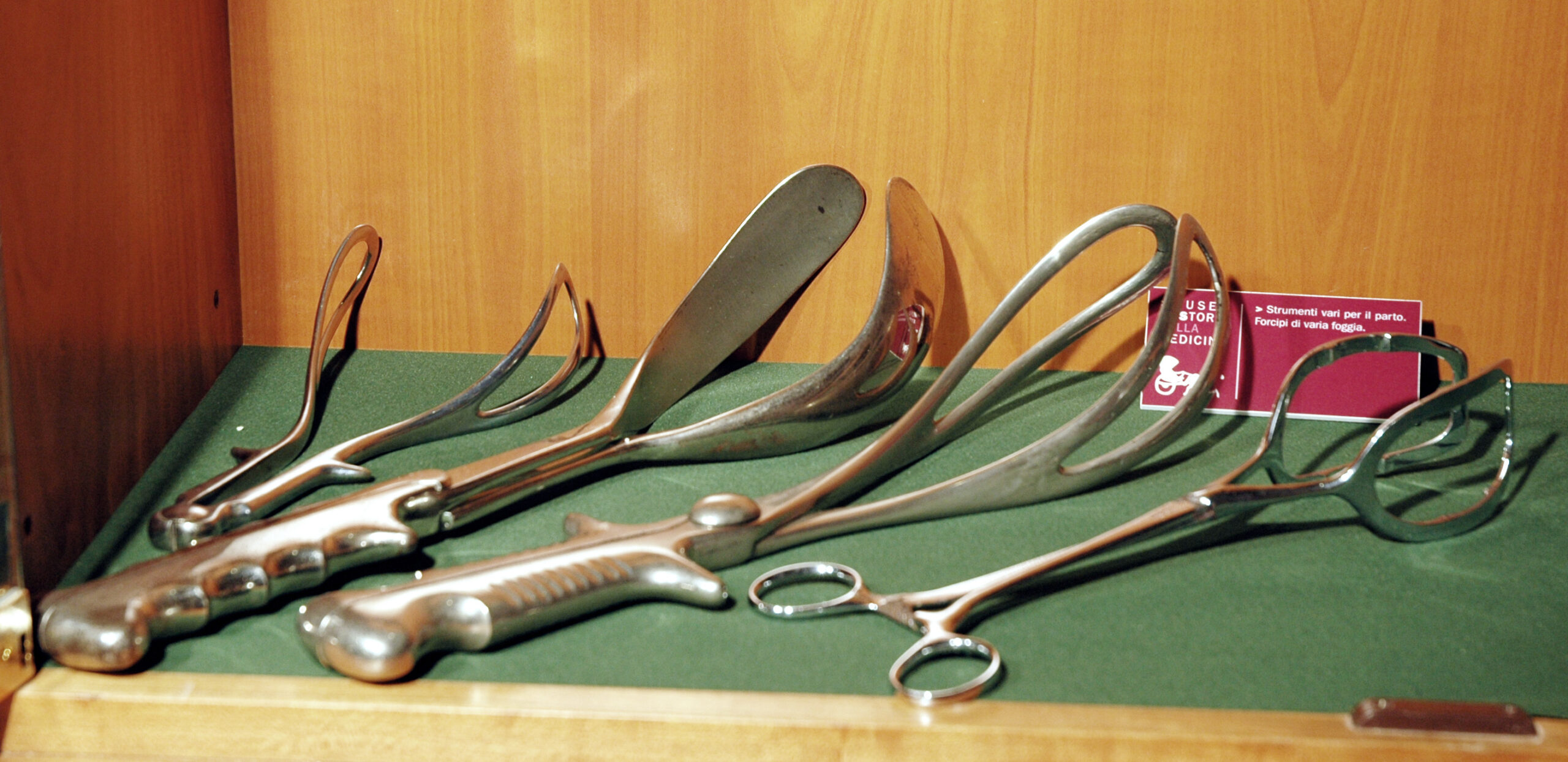 Ein Satz historischer geburtshilflicher Instrumente aus Metall, darunter Zangen und andere Instrumente, ausgestellt auf einem grünen Tuch in einer Holzkiste.